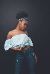Young slim African American female model with Afro hair bun and headband wearing stylish blue crop top and jeans standing against black background - ADSF21830