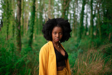 Junge Afroamerikanerin mit lockigem Haar, die einen eleganten leuchtend gelben Blazer trägt und in die Kamera schaut, während sie vor grünen Bäumen im Sommerwald steht - ADSF21835