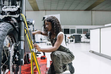 Seitenansicht einer afroamerikanischen Mechanikerin mit Schraubenschlüssel, die ein Motorrad repariert, während sie in der Werkstatt arbeitet - ADSF21837