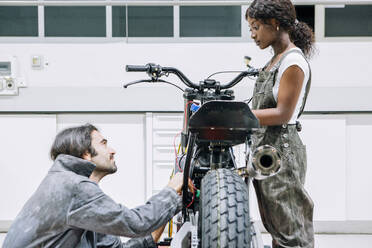 Diverse männliche und weibliche Mechaniker in Arbeitskleidung reparieren gemeinsam ein Motorrad in einer hellen Garage - ADSF21839