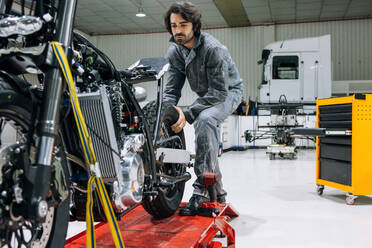 Seriöser männlicher Mechaniker in Arbeitskleidung, der in einer geräumigen Garage das Rad eines speziellen Motorrads repariert - ADSF21841