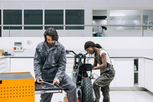 Beschäftigte multiethnische Männer und Frauen in schmutziger Arbeitskleidung reparieren ein Motorrad und benutzen dabei Instrumente aus einem Werkzeugschrank - ADSF21846