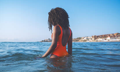 Seitenansicht einer nicht erkennbaren afroamerikanischen Touristin mit lockigem Haar in einem schicken roten Badeanzug, die im wogenden Meer vor einem wolkenlosen blauen Himmel steht - ADSF21849