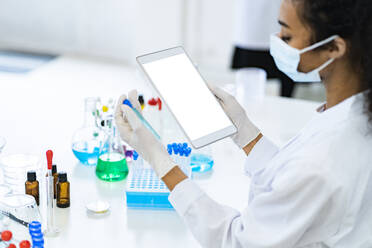 Junger Forscher, der ein Reagenzglas untersucht, während er ein digitales Tablet im Labor während einer Pandemie hält - GIOF11863
