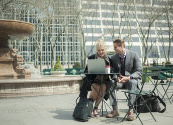 Smiling business couple with laptop sitting in public park - AJOF01273