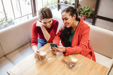 Lächelnde Frau, die mit ihrer Freundin im Café ein Mobiltelefon benutzt - LJF02128