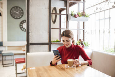 Lächelnde Frau, die ein Mobiltelefon benutzt, während sie einen Kaffee im Café trinkt - LJF02131