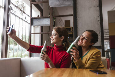 Cheerful female friends with beer bottles taking selfie through mobile phone in bar - LJF02145