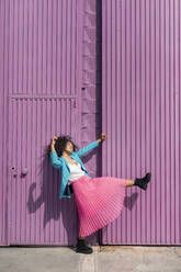 Verspielte junge Frau tanzt an einer lila Hüttenwand an einem sonnigen Tag - TCEF01679