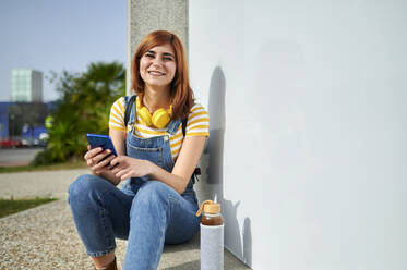 Glückliche junge Frau sitzt mit Smartphone an einer weißen Wand an einem sonnigen Tag - KIJF03636