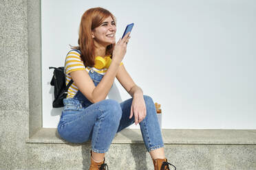 Fröhliche rothaarige Frau, die über einen Lautsprecher mit ihrem Smartphone spricht, während sie vor einer weißen Wand sitzt - KIJF03637