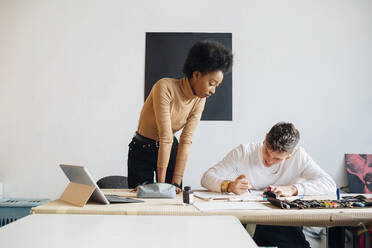 Young woman looking at man writing with fountain pen while sitting at table in studio - MEUF02107