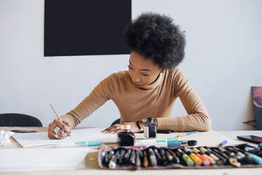 Afro woman writing on paper with pen while sitting at table in studio - MEUF02126