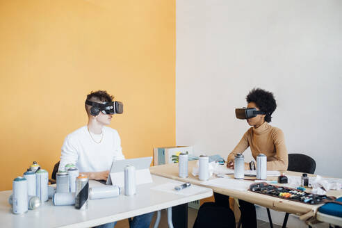 Young multi ethnic couple wearing virtual reality simulator sitting at table in studio - MEUF02147