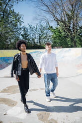 Multi ethnic couple walking while holding hands at skateboard park during sunny day - MEUF02152