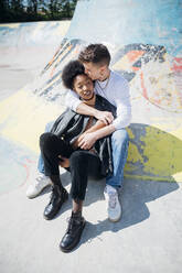 Young man kissing on girlfriend's forehead while sitting at skateboard park during sunny day - MEUF02153