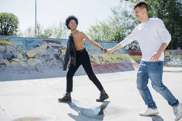 Verspieltes junges Paar, das sich in einem Skateboard-Park an einem sonnigen Tag an den Händen hält - MEUF02156