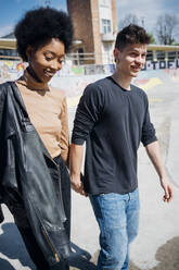 Lächelndes multiethnisches Paar, das in einem Skateboard-Park an einem sonnigen Tag spazieren geht und die Hände hält - MEUF02158