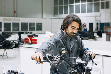 Fokussierter männlicher Mechaniker in Uniform, der den Lenker eines Motorrads untersucht, während er in einer geräumigen Werkstatt arbeitet - ADSF21850