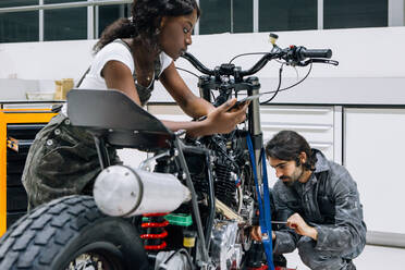 Schwarze Mechanikerin mit Tablet und männlicher Techniker, der mit einem Schraubenzieher ein Motorradrad repariert, während sie gemeinsam in einer geräumigen Werkstatt arbeiten - ADSF21855