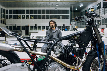 Adult bearded male technician sitting near disassembled custom motorbike while working in modern spacious workshop - ADSF21863