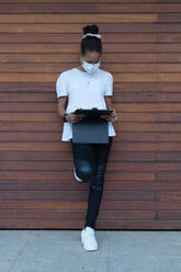 Full body of unrecognizable young African American female in casual outfit and medical mask leaning on wooden wall and using tablet on street during coronavirus - ADSF21880