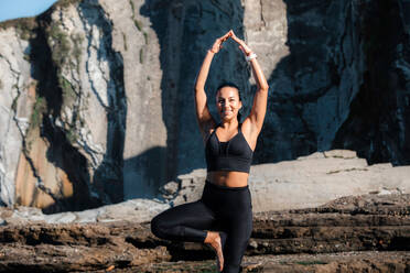 Flexible female in sportswear balancing on leg in Vrksasana with raised arms while doing yoga in mountains in summer - ADSF21887