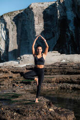 Flexible Frau in Sportkleidung balanciert auf einem Bein in Vrksasana mit erhobenen Armen beim Yoga in den Bergen im Sommer - ADSF21888