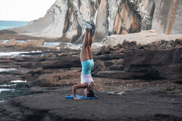 Rückenansicht einer ruhigen Frau, die in Salamba Sirsasana am felsigen Strand balanciert, während sie im Sommer Yoga übt - ADSF21891