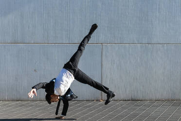 Asiatischer männlicher Unternehmer in formellem Anzug zeigt Breakdance-Bewegung und balanciert auf dem Arm im Handstand auf einer Straße in der Stadt, während er in die Kamera schaut - ADSF21905
