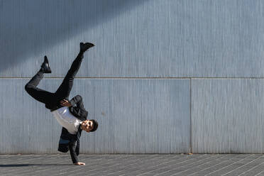 Asiatischer männlicher Unternehmer in formellem Anzug zeigt Breakdance-Bewegung und balanciert auf dem Arm im Handstand auf einer Straße in der Stadt, während er in die Kamera schaut - ADSF21906