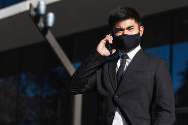 Side view of Asian businessman in protective mask and elegant suit standing in city and having smartphone conversation while looking away - ADSF21914