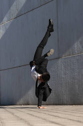 Asiatischer männlicher Unternehmer in formellem Anzug zeigt Breakdance-Bewegung und balanciert auf dem Arm im Handstand auf einer Straße in der Stadt, während er in die Kamera schaut - ADSF21915