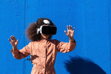 Unerkanntes afroamerikanisches Mädchen in modischem Anzug und VR-Headset, das die Luft berührt, während es die virtuelle Welt vor einer blauen Wand auf der Straße erkundet - ADSF21923