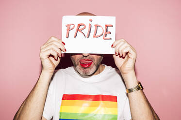 Rebellious bearded homosexual man with red lips and manicure making grimace with tongue out while showing and covering face with paper with Pride text against pink background - ADSF21928