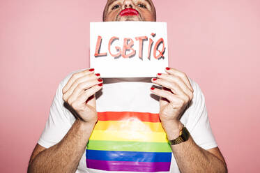 Rebellious bearded homosexual man with red lips and manicure making grimace while showing and covering face with paper with LGBTIQ text against pink background - ADSF21929