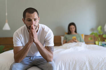 Worried young man sitting on bed while girlfriend using smart phone in background at home - SBOF03178