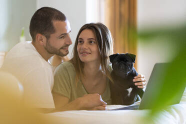 Junge Frau mit Mops starrt ihren Freund an, der auf den Laptop schaut, während sie zu Hause auf dem Bett liegt - SBOF03191