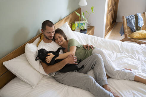 Playful couple embracing Pug Dog while lying on bed at home - SBOF03194