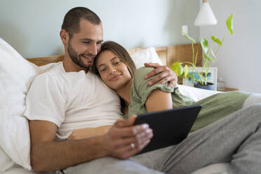 Lächelndes junges Paar, das ein digitales Tablet benutzt, während es zu Hause auf dem Bett liegt - SBOF03197