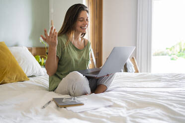 Young woman gesturing during video call on laptop while sitting on bed at home - SBOF03200