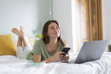 Nachdenkliche junge Frau mit Smartphone, die zu Hause auf dem Bett vor dem Laptop liegt - SBOF03202