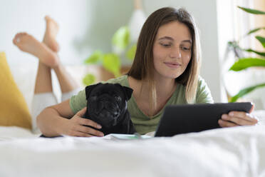 Junge Frau umarmt Mops, während sie zu Hause ein digitales Tablet benutzt - SBOF03205