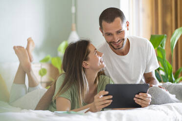 Junge Frau mit digitalem Tablet, die ihren Mann zu Hause auf dem Bett sitzend betrachtet - SBOF03206