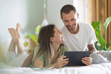 Young woman with digital tablet looking at husband sitting on bed at home - SBOF03206