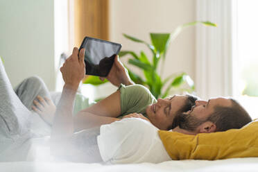 Junges Paar benutzt ein digitales Tablet, während es zu Hause auf dem Bett liegt - SBOF03212