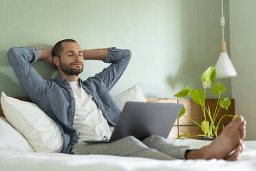 Relaxed man sitting with laptop on lap in bedroom at home - SBOF03218