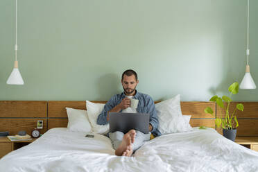 Young man having coffee using laptop while sitting on bed at home - SBOF03220