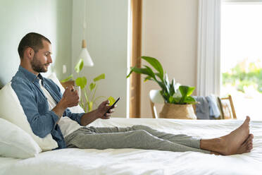 Junger Mann mit Kaffeetasse und Smartphone zu Hause - SBOF03222