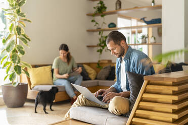 Young man working on laptop while wife looking at Pug dog in background at home - SBOF03225